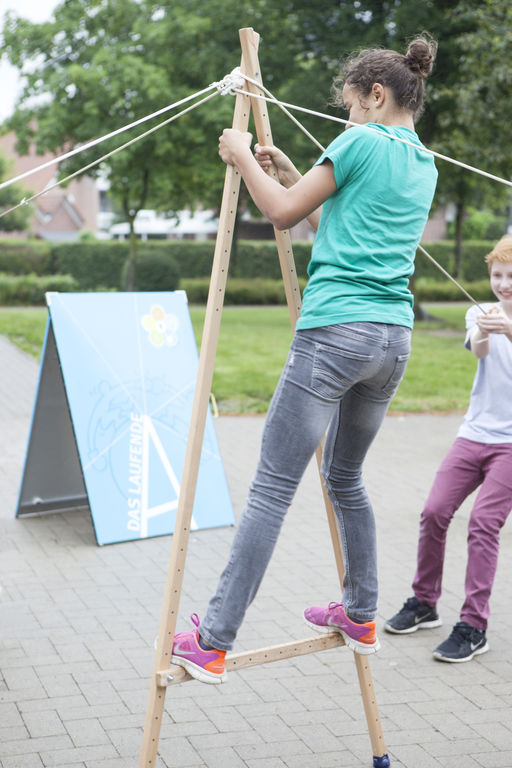 Ein Kind balanciert auf einem hölzernen A, während mehrere Mitschüler*innen die Konstruktion mit Seilen stabilisieren 
