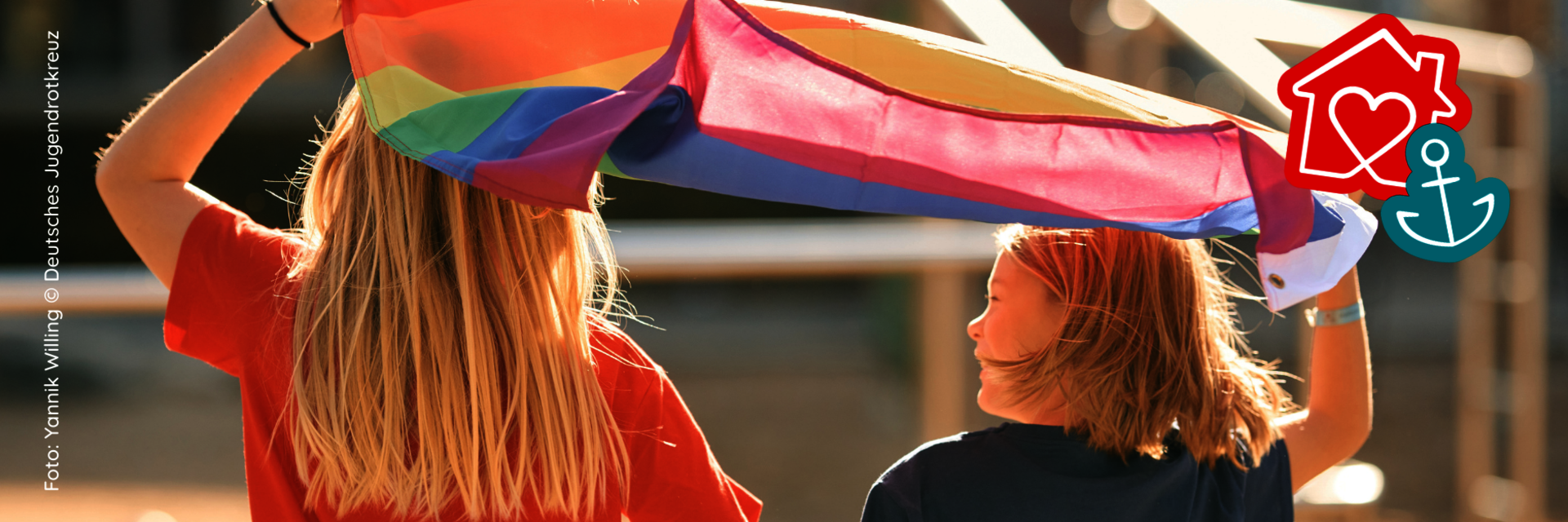 Zwei Kinder stehen mit dem Rücken zu uns. Sie halten gemeinsam eine Pride-Flagge über ihren Kopf. Sie weht im Wind.