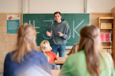 Ein JRKler steht in einem Klassenraum vorne an der Tafel Ein junger Erwachsener steht in einem Klassenzimmer vor einer Tafel auf der JRK steht