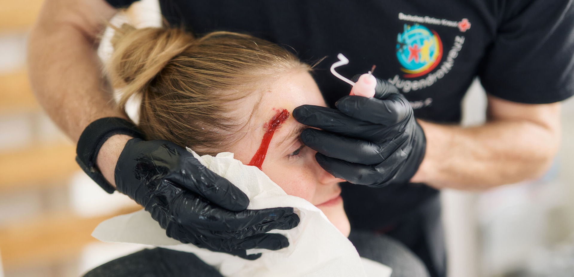 Notfalldarstellung: Helfer vom JRK beim Schminken einer Wunde am Kopf einer Darstellerin für ein Erste Hilfe 